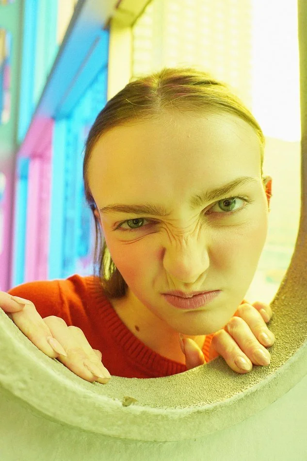Sadie, a young Montréal female model signed with MTL Models MGMT with brown hair and green eyes making a funny face, squinting one eye and wrinkling her nose, looking through a round opening.