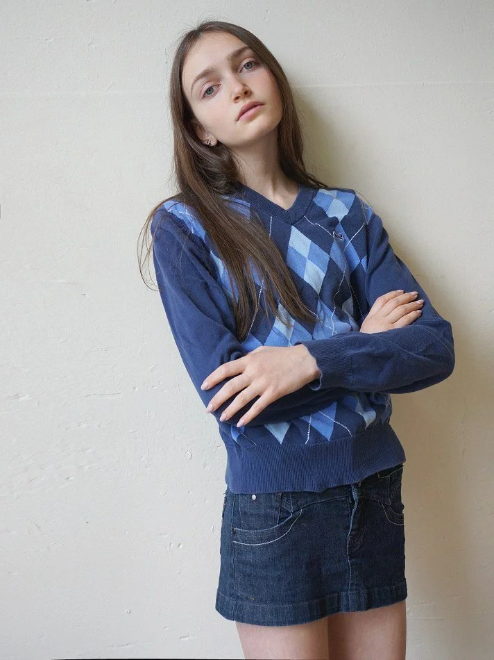 Sadie, a young Montréal female model signed with MTL Models MGMT with long brown hair modelling against a plain off-white wall, crossing her arms, wearing a blue argyle sweater and a dark denim skirt in a Montreal studio.