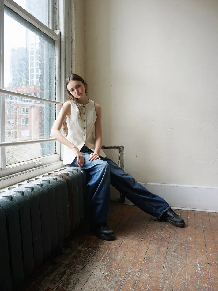 Sadie, a young Montréal female model signed with MTL Models MGM with long brown hair sitting on a window sill in a room with white walls and wooden floor. She is wearing a sleeveless beige top and loose-fitting dark blue jeans, looking at the camera.