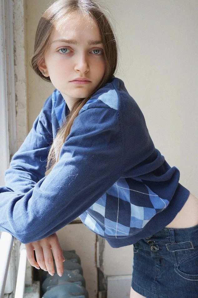 Sadie, a young Montréal female model signed with MTL Models MGMT with long, straight hair and blue eyes, wearing a blue argyle sweater and dark shorts, leaning on a white railing near a window in a well-lit room.
