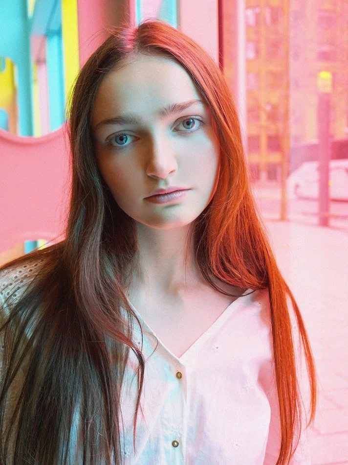 Sadie, a young Montréal female model signed with MTL Models MGMT with long brown hair and blue eyes stands indoors, with colorful vertical blinds and a window showing an urban street scene in the background.