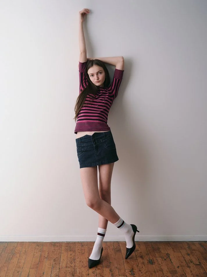 Sadie, a young Montréal female model signed with MTL Models MGMT with long brown hair, wearing a pink and black striped top, a denim skirt, white socks, and black high heels, modelling against a plain white wall with one arm raised above her head.