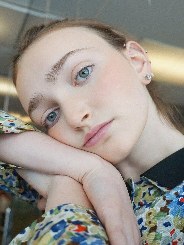Close-up of model Sadie modelling her head on her arm, with her face turned slightly to the side, looking into the camera. She has blue eyes, light makeup, and multiple earrings. Shot by Montreal photographer Melina Querel.