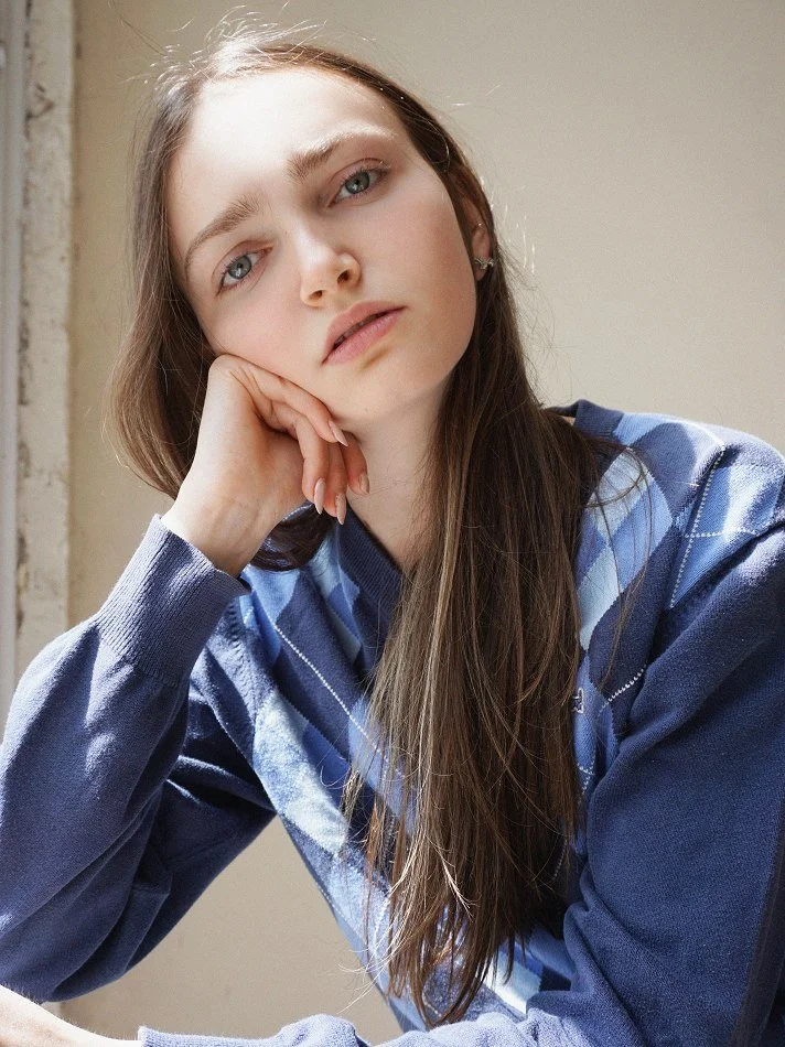 Sadie, a young Montréal female model signed with MTL Models MGMT with long brown hair and blue eyes resting her head on her hand, wearing a blue plaid shirt, sitting near a window with sunlight.  Shot by photographer Melina Querel.