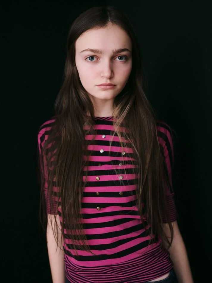 Sadie, a young Montréal female model signed with MTL Models MGMT with long brown hair and blue eyes wearing a pink and black striped shirt with gold embellishments against a black background.