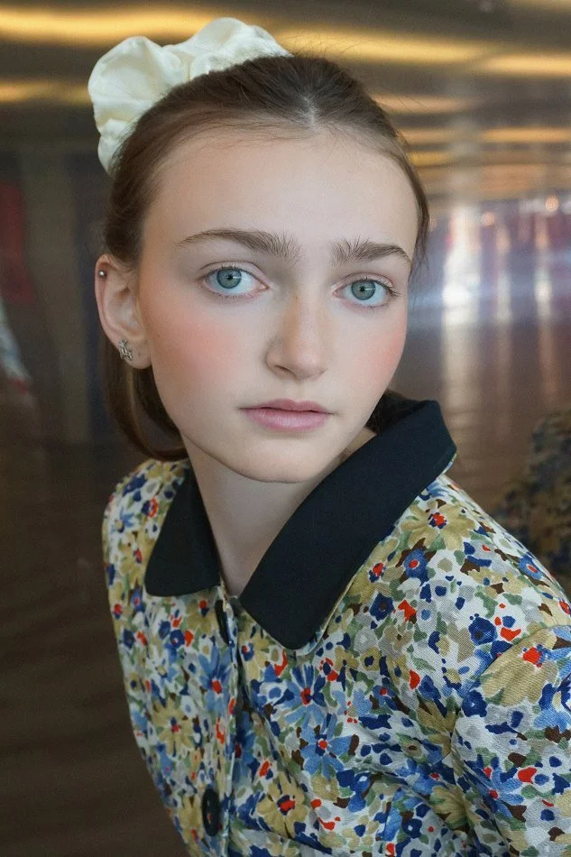 Sadie, a young Montréal female model signed with MTL Models MGMT with light skin and bright blue eyes, wearing a floral-patterned shirt and a white headband, modelling indoors.