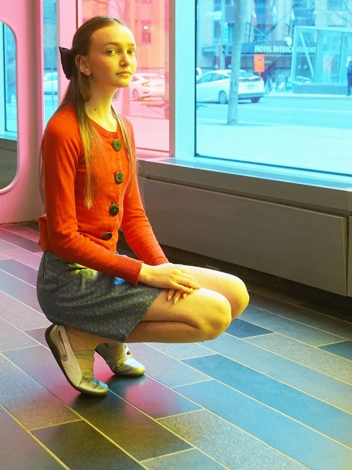 Sadie, model with long, light brown hair tied with a black bow, wearing a red cardigan with large buttons and a blue skirt with orange polka dots, squatting on the floor inside a building near large glass windows showing a city street outside.