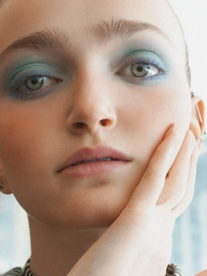 Close-up of Sadie, a young Montréal female model signed with MTL Models MGMT with blue eyes and pastel-colored makeup on her eyelids, modelling touching her face with her right hand.