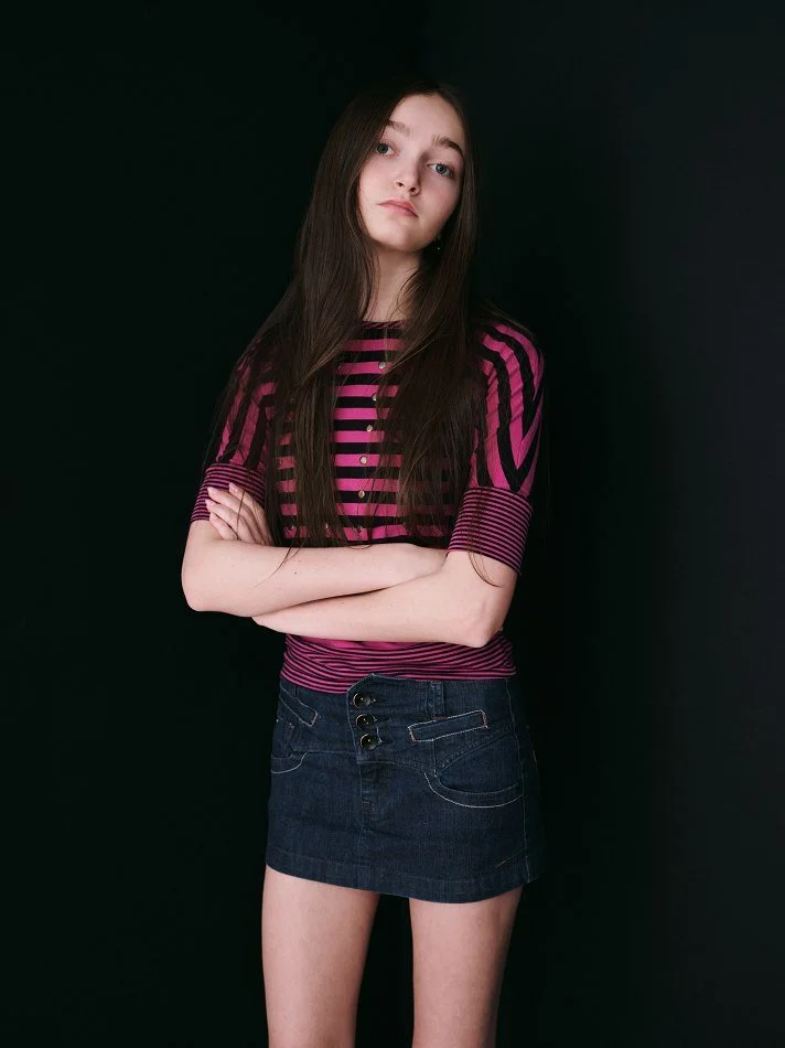 Sadie, a young Montréal female model signed with MTL Models MGMT with long brown hair wearing a pink and purple striped t-shirt and dark denim skirt, standing against a black background with arms crossed.