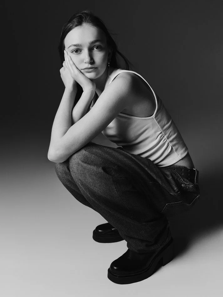 Sadie, a young Montréal female model signed with MTL Models MGMT crouching down against a dark background, resting her face on her hand, looking directly at the camera, wearing a sleeveless top, loose jeans, and black boots in black and white.