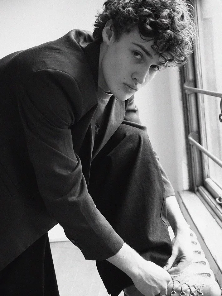 Albert, Montréal male model signed with MTL Models MGMT with curly hair sitting by a window, looking at the camera, dressed in a suit, in a black and white photo.