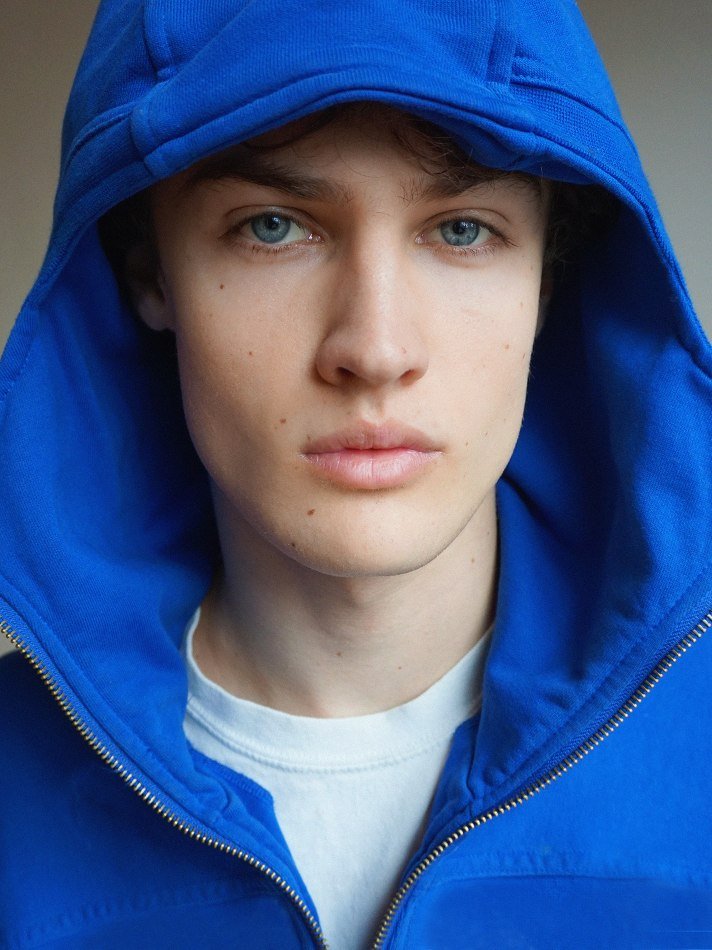 Albert, Montréal male model signed with MTL Models MGMT with blue eyes wearing a blue hoodie with the hood up, facing the camera directly.