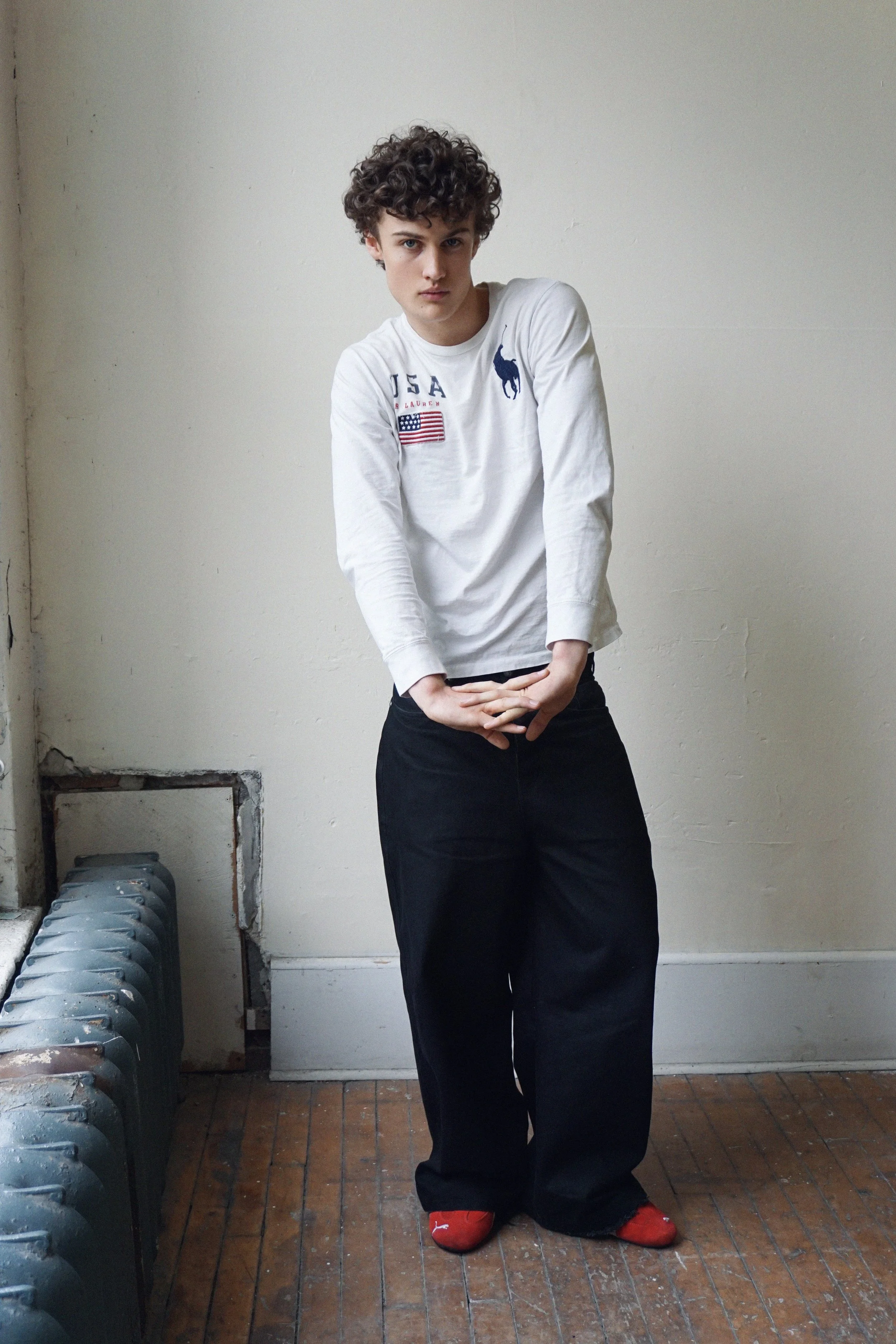 Albert, Montréal male model signed with MTL Model with curly hair standing indoors against a plain white wall, wearing a white long sleeve shirt with logos, black wide-leg pants, and red socks with white Puma logo, posing with hands clasped in front.