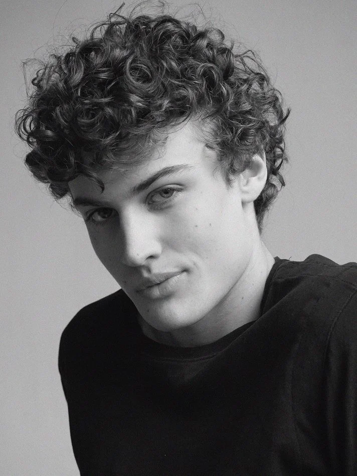 Black and white portrait of Albert, Montréal male model signed with MTL Models MGMT with curly hair, looking at the camera with a slight smile, wearing a dark shirt.