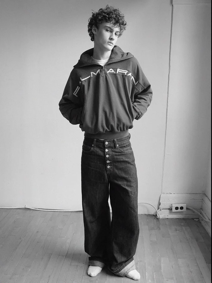 Albert, Montréal male model signed with MTL Models MGMT with curly hair standing against a plain wall, wearing a loose hoodie, wide-legged jeans, and socks, looking at the camera.