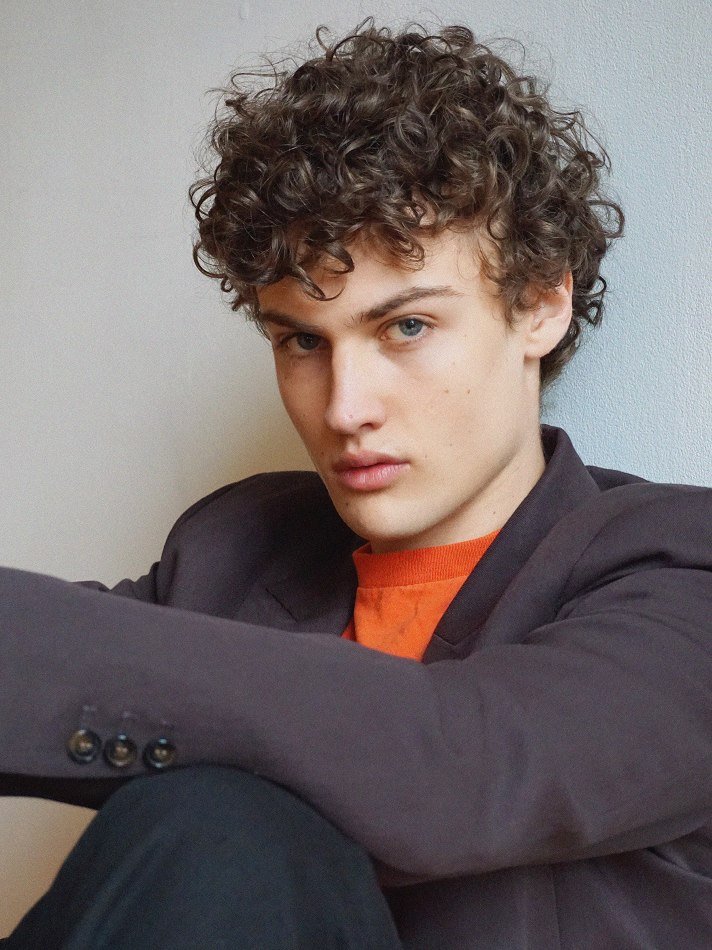 Albert, Montréal male model signed with MTL Models MGMT with curly brown hair and blue eyes, wearing a dark blazer over an orange shirt, sitting against a light-colored wall.