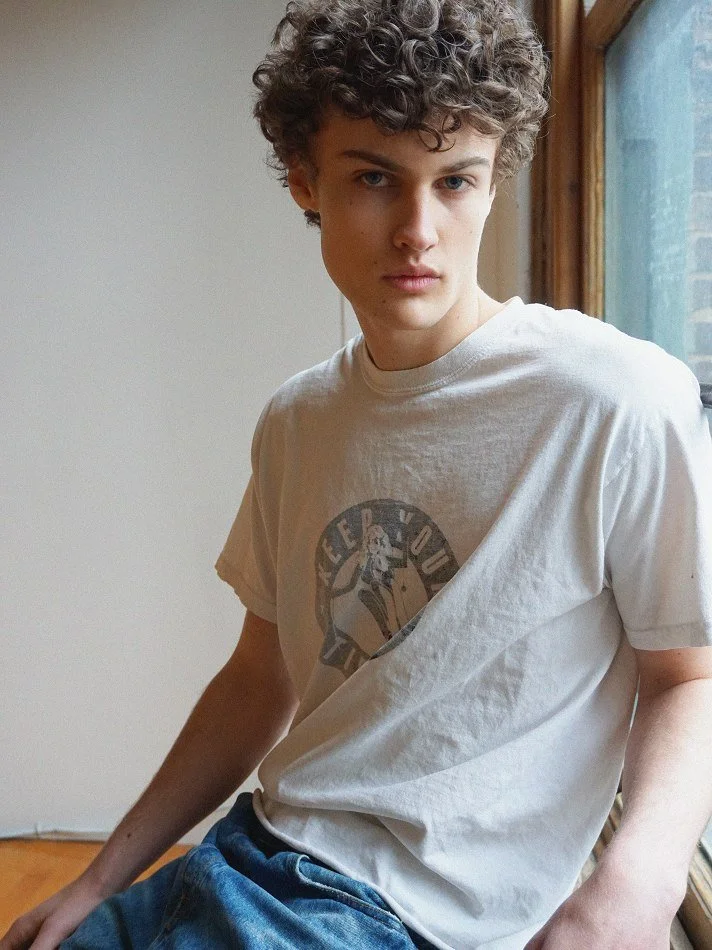 Albert, Montréal male model signed with MTL Models MGMT with curly hair wearing a graphic t-shirt and jeans, sitting near a window.