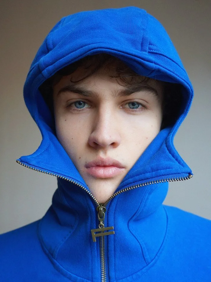 Albert, Montréal male model signed with MTL Models MGMT with blue eyes wearing a bright blue hoodie with the hood up, zipping up the front, against a plain neutral background.