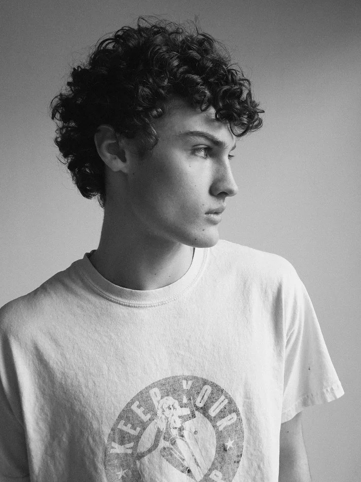 Black and white photo of Albert, Montréal male model signed with MTL Models MGMT with curly hair, wearing a T-shirt with a vintage-style logo or graphic on the front, looking to the side.
