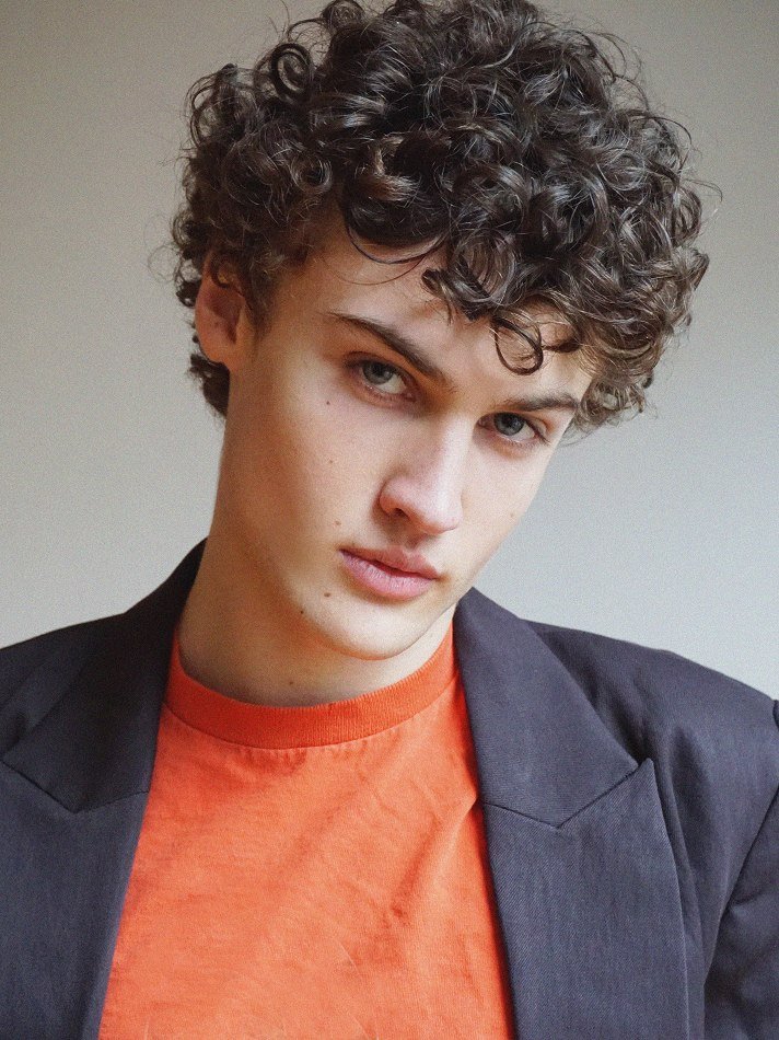 Albert, Montréal male model signed with MTL Models MGMT with curly brown hair, blue eyes, wearing an orange t-shirt and a dark blazer, looks into the camera with a slight smirk against a plain background.