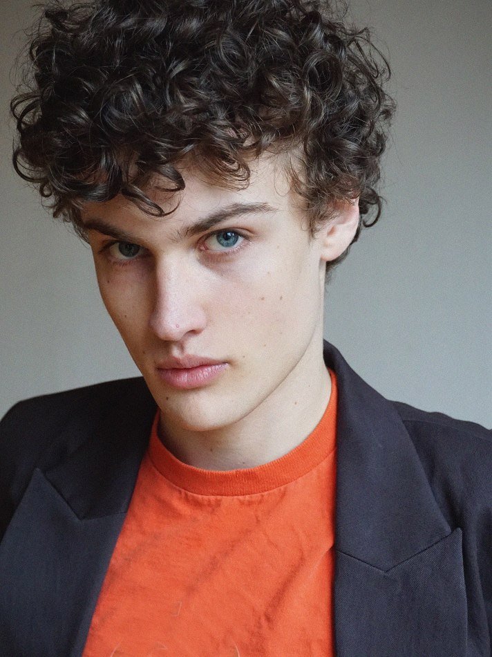 Close-up portrait of Albert, Montréal male model signed with MTL Models MGMT with curly brown hair and blue eyes, wearing an orange t-shirt and a black blazer.