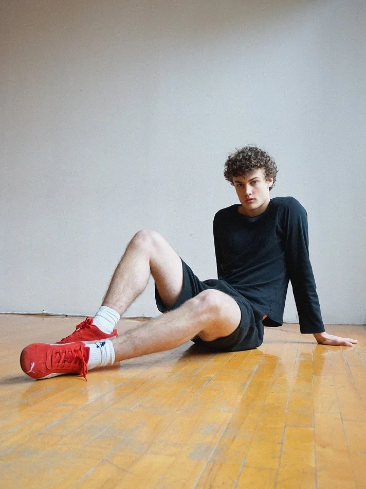 Albert, Montréal male model signed with MTL Models MGMT sitting on wooden floor with one leg extended and the other bent, wearing black long sleeve shirt, black shorts, white socks, and red sneakers, against a plain white wall.