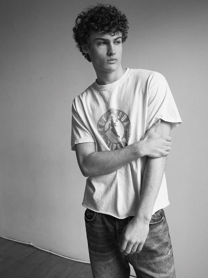 Albert, Montréal male model signed with MTL Models MGMT with curly hair standing against a plain wall, wearing a T-shirt and jeans, looking to the side in a contemplative pose.