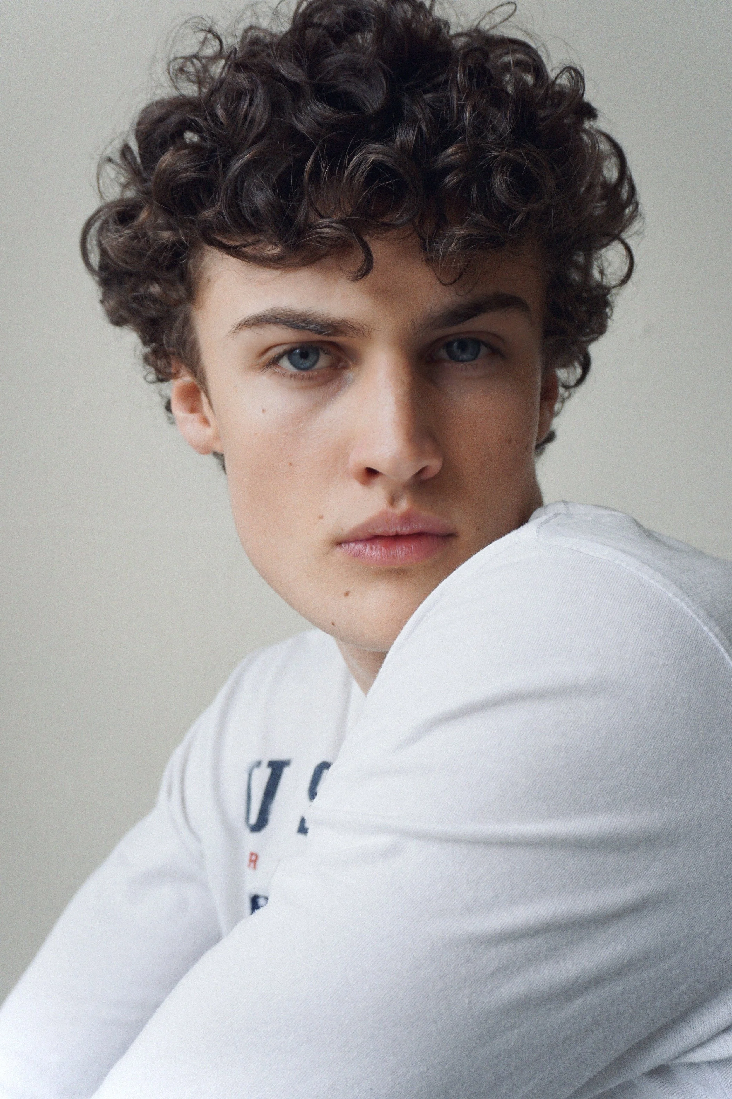 Close-up of Albert, Montréal male model signed with MTL Models MGMT with short, curly dark hair, blue eyes, and light skin, wearing a white shirt with text partially visible, against a plain neutral background.