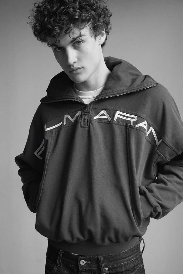 Albert, Montréal male model signed with MTL Models MGMT with curly hair wearing a zip-up jacket with the brand name  across the chest, posing with hands in pockets against a plain background.