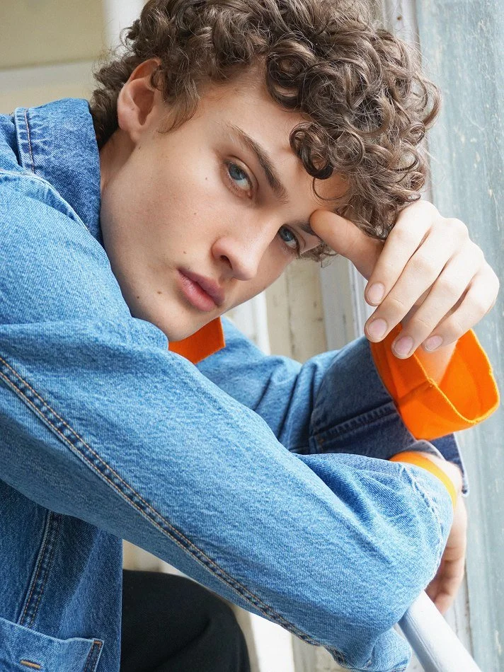 Albert, Montréal male model signed with MTL Models MGMT with curly hair and light skin is leaning on his arm, looking directly at the camera with a serious expression. He is wearing a denim jacket with orange accents and sitting near a window.