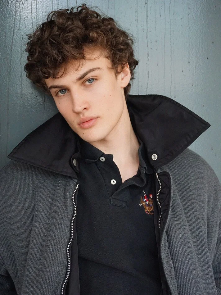 Albert, Montréal male model signed with MTL Models MGMT with curly hair and blue eyes leaning against a gray wall, wearing a black polo shirt with a crest, a gray jacket, and a black collar.