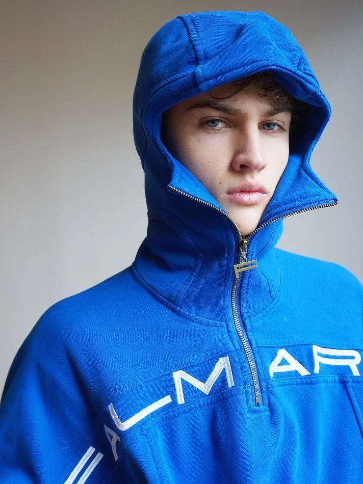 Albert, Montréal male model signed with MTL Models MGMT wearing a blue hooded jacket with the zipper partially open, standing against a plain background.