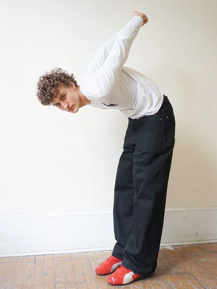 Albert, Montréal male model signed with MTL Models MGMT with curly hair wearing a white long-sleeve shirt, dark pants, and red sneakers, bending forward with arms raised behind back, against a plain off-white wall.