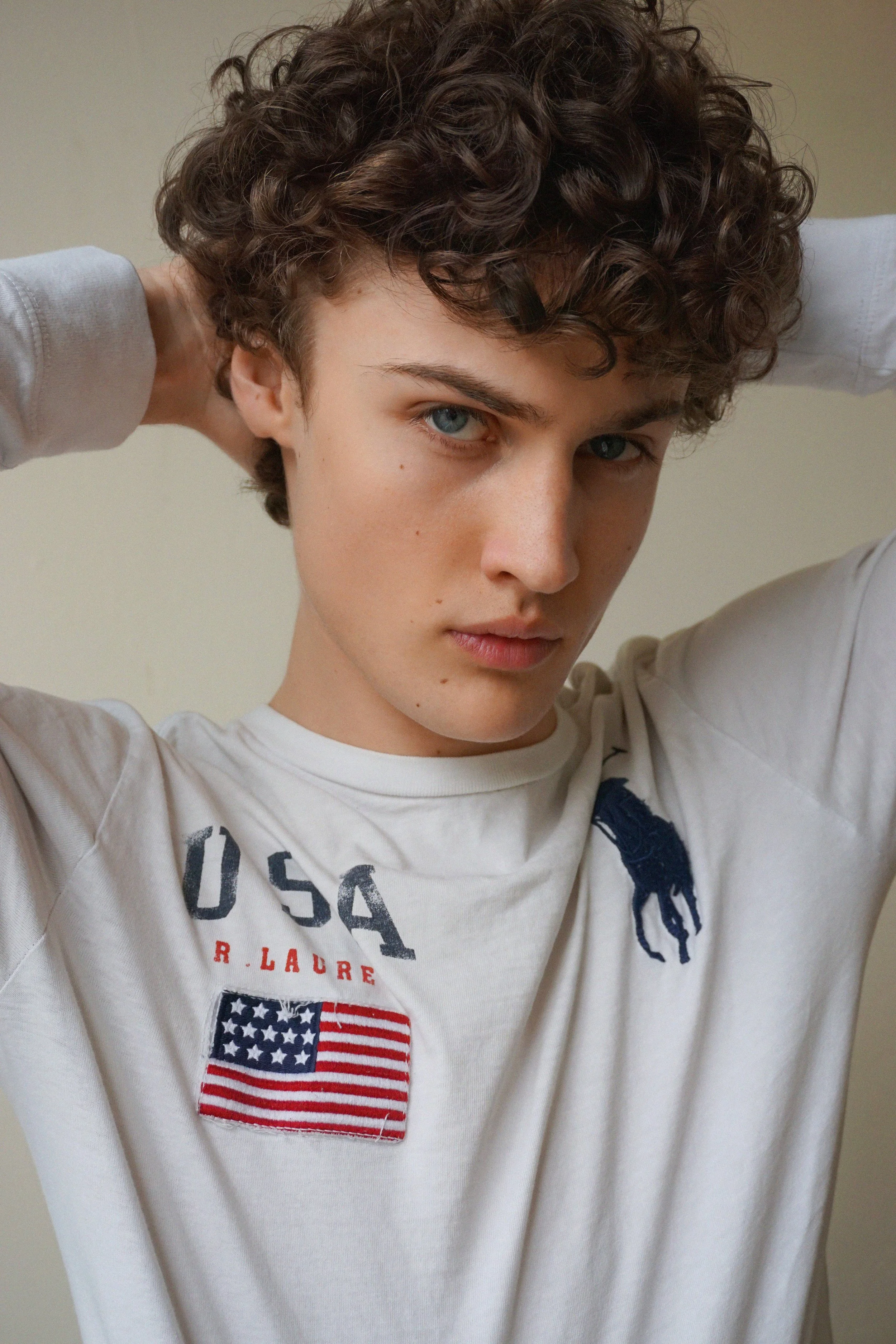 Albert, Montréal male model signed with MTL Models MGMT with curly brown hair and blue eyes is looking at the camera and holding their hands on their head. They are wearing a white USA-themed sweatshirt featuring an American flag patch.