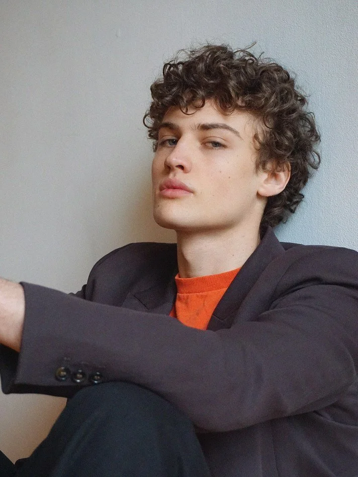 Young man with curly brown hair wearing a blazer and orange shirt, sitting against a plain wall, looking confidently at the camera.