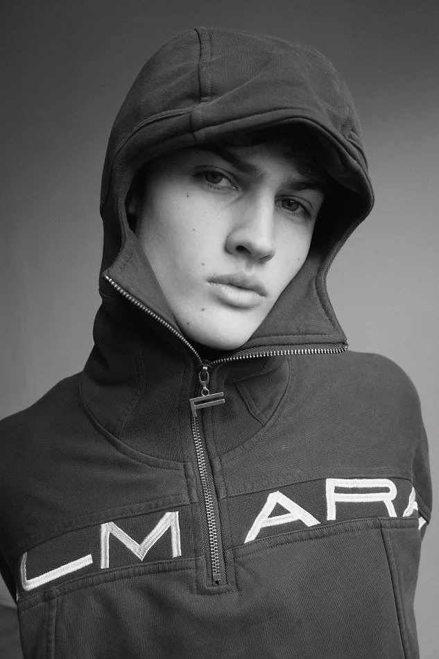 Albert, Montréal male model signed with MTL Models MGMT taking a portrait wearing a hooded jacket with the zipper partially open, and the brand name "CAMEO" visible on the chest, in black and white.