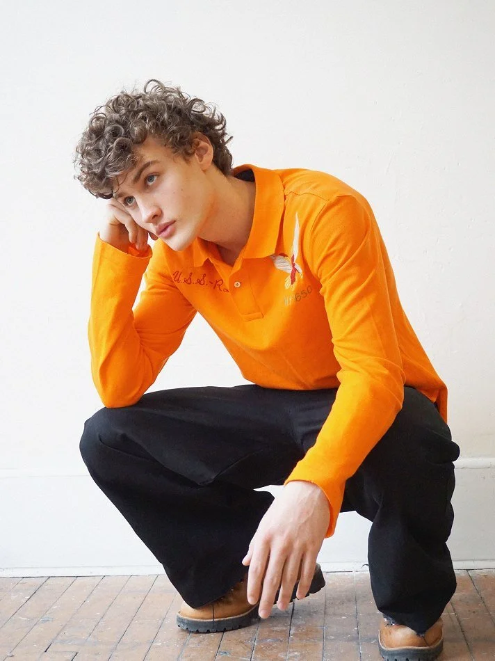 Albert, Montréal male model signed with MTL Models MGMT with curly hair wearing an orange long-sleeve shirt and black pants, squatting indoors with a contemplative expression.