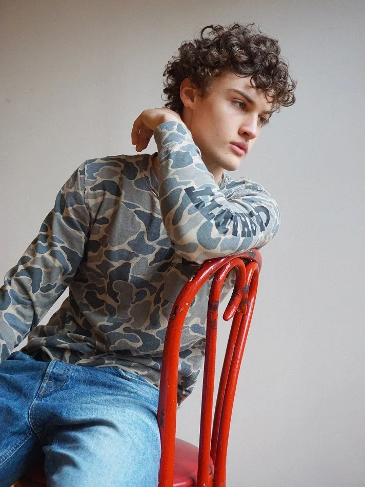 Albert, Montréal male model signed with MTL Models MGMT with curly hair sitting on a red chair, wearing a camouflage long sleeve shirt and blue jeans, with his arm resting on the backung man of the chair and looking contemplative.