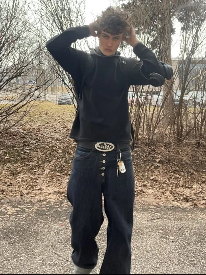 A young person with curly hair adjusting their hair outdoors, wearing a black hoodie, baggy dark jeans with button details, a belt with a large oval buckle, and headphones around their neck, in a wooded area with leafless trees and parked cars in the