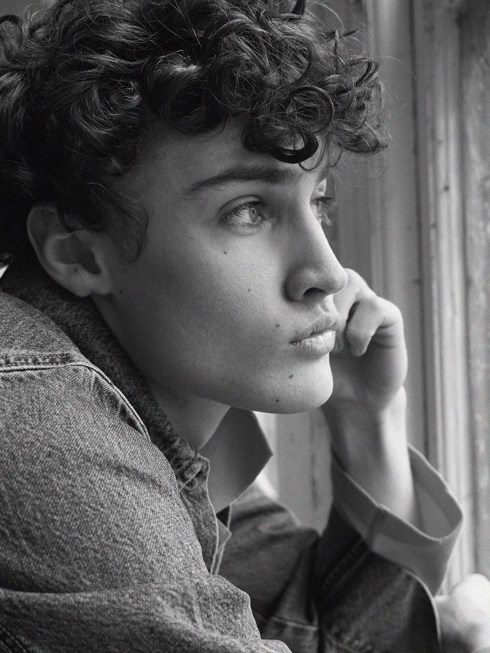 Black and white photo of a young man with curly hair looking out a window in deep thought, resting his chin on his hand.
