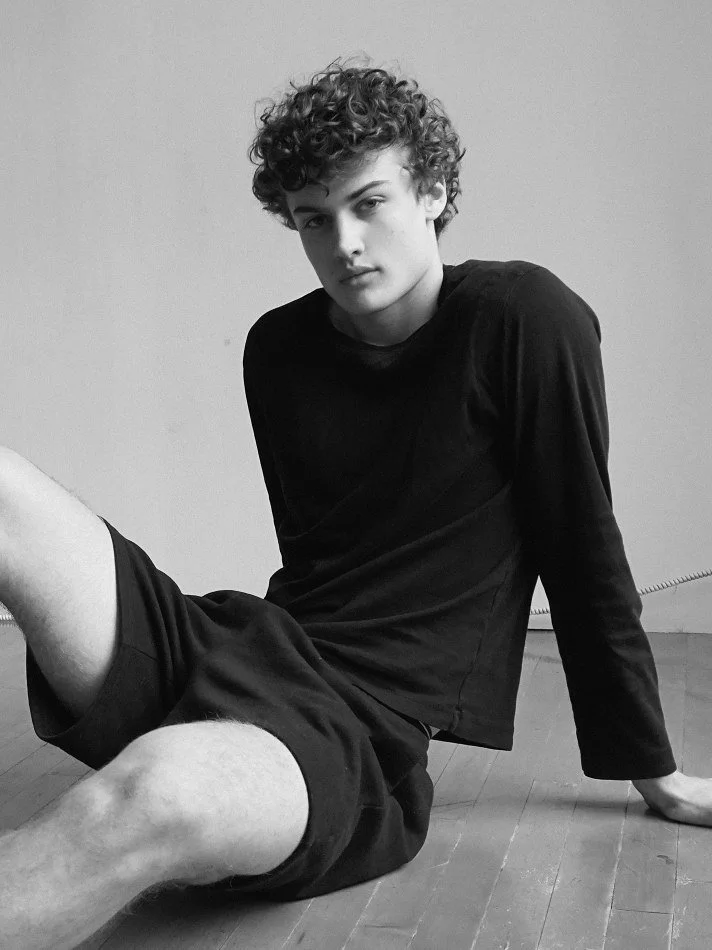Black and white photo of Albert, Montréal male model signed with MTL Models MGMT with curly hair sitting on the floor, wearing a black long-sleeve shirt and shorts, with a plain wall background.