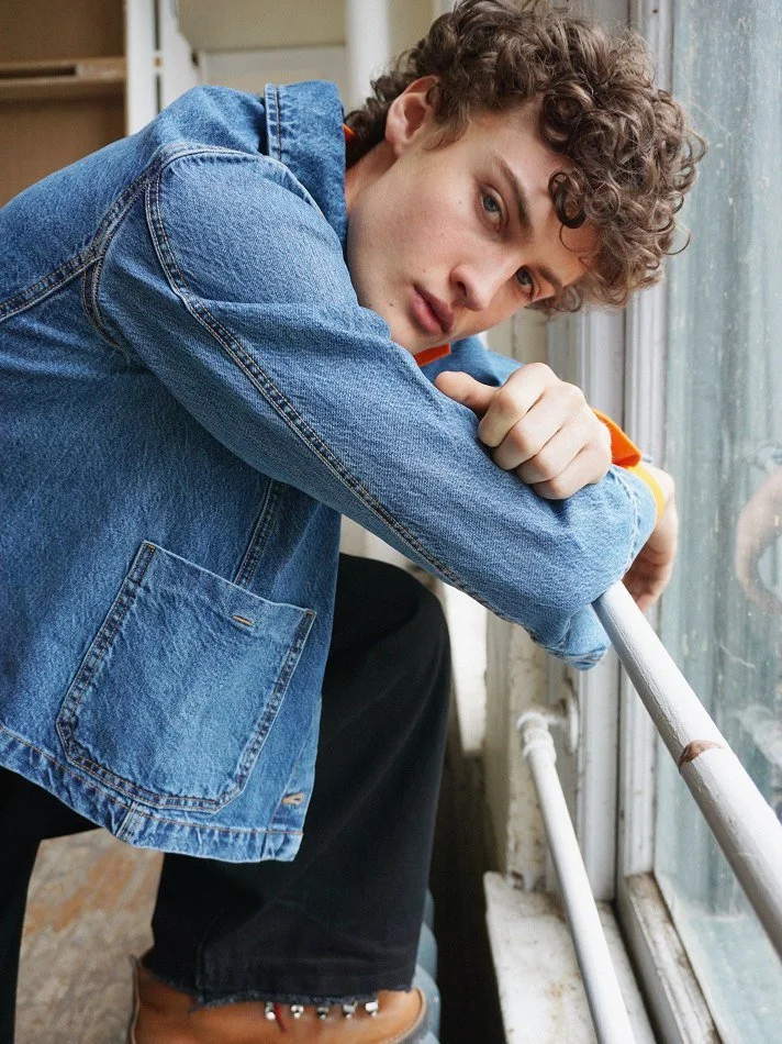 A young person with curly hair, wearing a denim jacket and black pants, leaning on a railing near a window, looking at the camera.