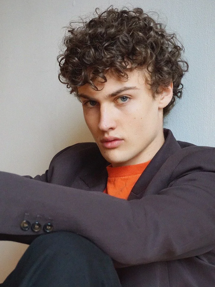 A young man with curly brown hair and blue eyes sitting against a plain light-colored wall, wearing a dark blazer and an orange shirt, looking serious at the camera.