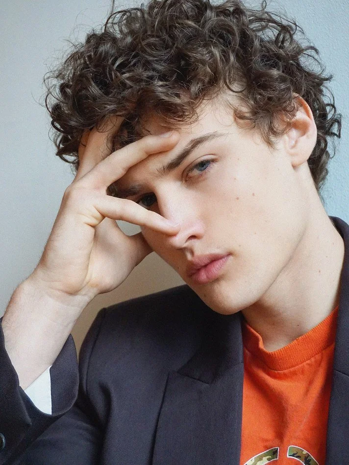 Close-up of a young man with curly hair, wearing a dark blazer over an orange shirt, resting his head on his hand with a neutral expression.