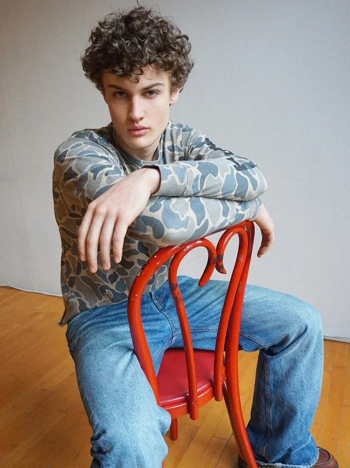 Young man with curly hair sitting on a red chair, leaning forward with one arm resting on the back of the chair, looking at the camera, wearing a camouflage-patterned long-sleeve shirt and blue jeans.
