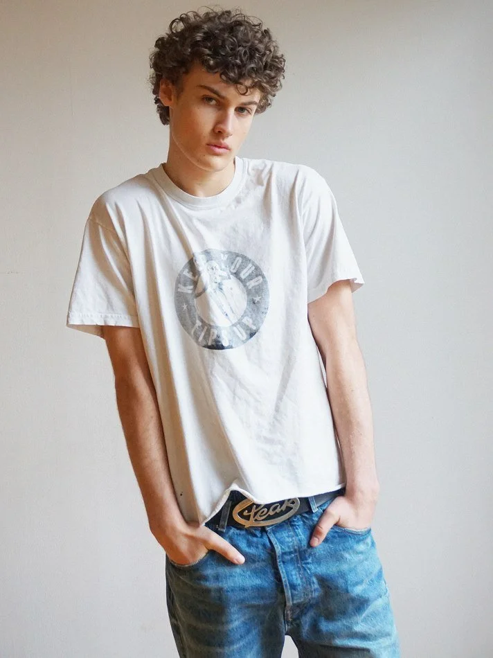 A young man with curly hair wearing a white graphic T-shirt and blue jeans, standing against a plain white wall.
