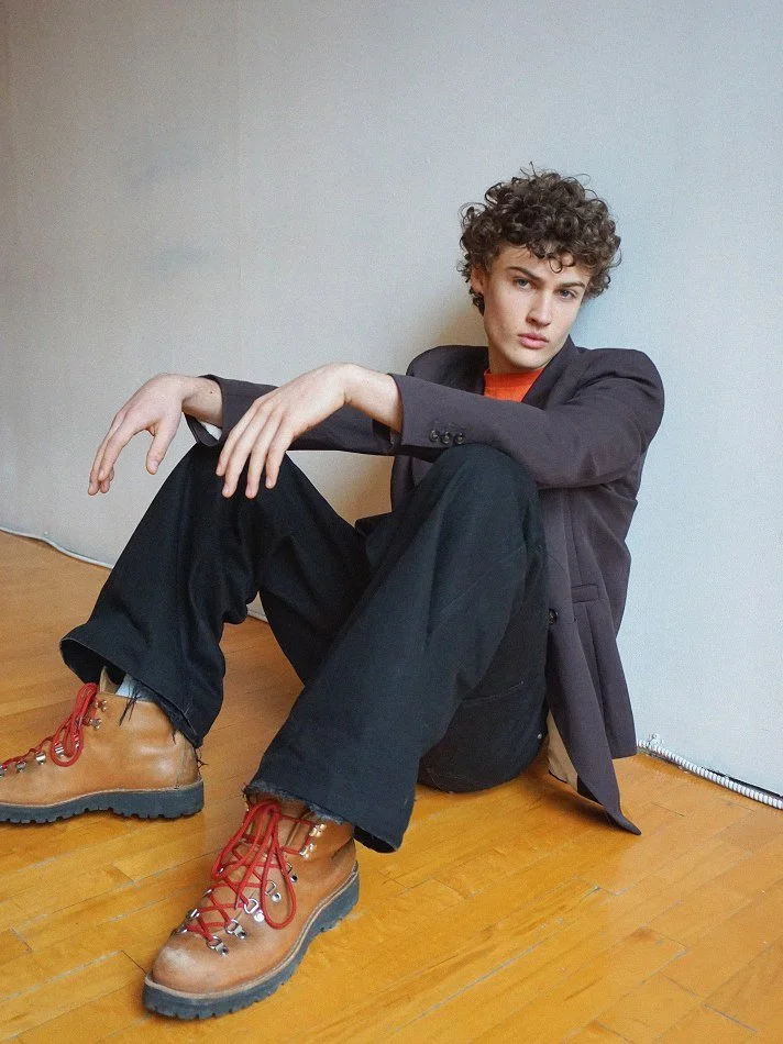 A young person with curly hair is sitting on a wooden floor, leaning against a wall. They are wearing a dark blazer, black pants, and tan hiking boots with red laces. They have a relaxed, introspective expression.