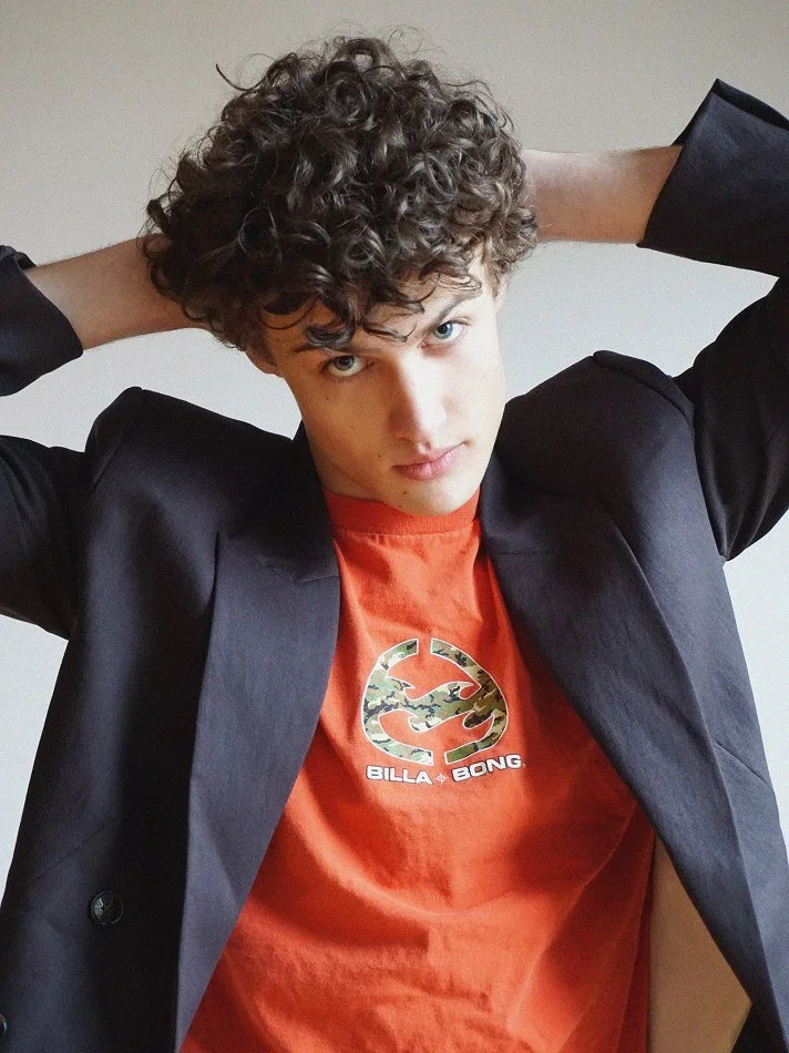 A young man with curly hair wearing a black blazer and a red BILLA BONG t-shirt, posing with his hands behind his head against a plain wall.