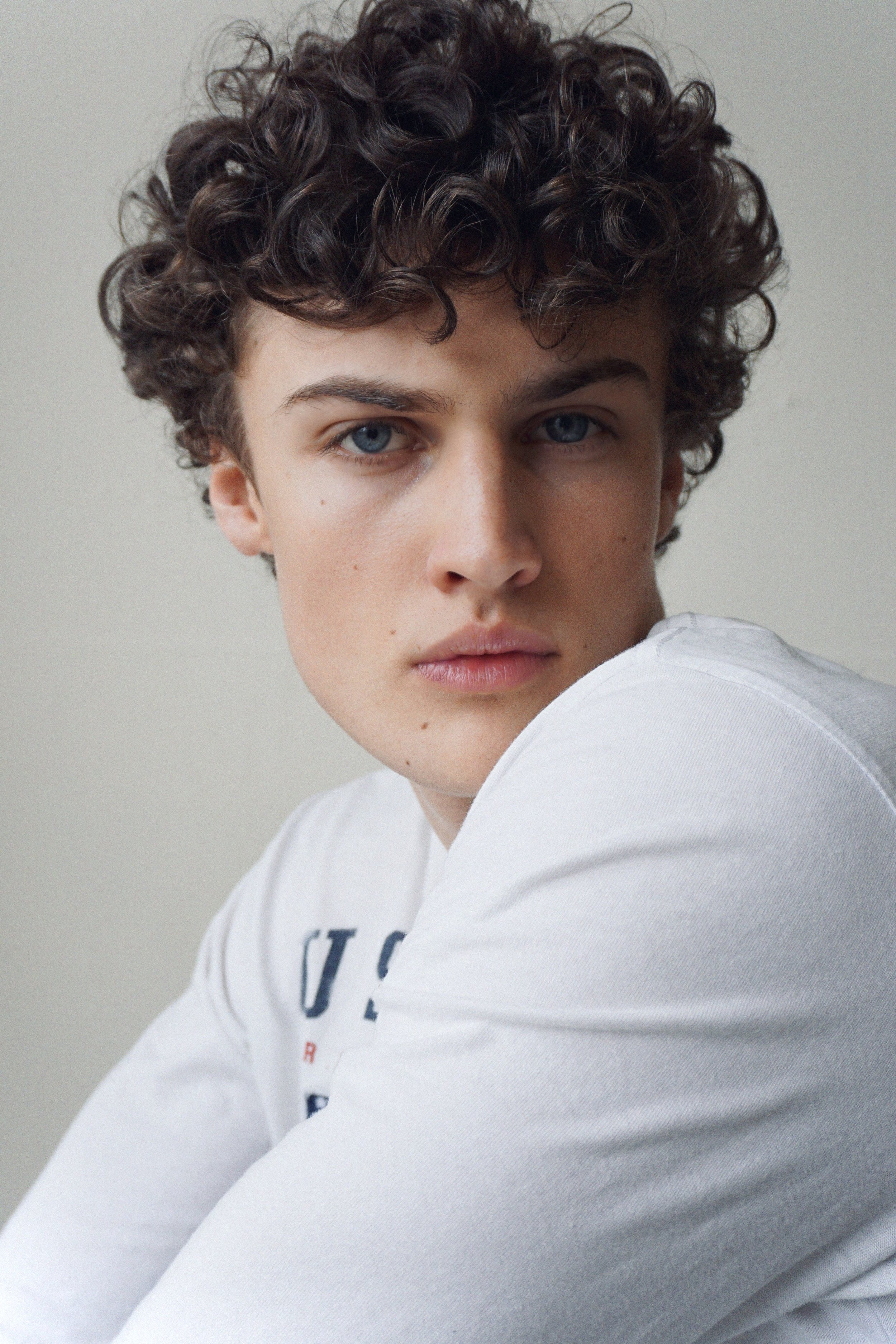 A young person with short, curly brown hair and blue eyes looking directly at the camera, wearing a white shirt.