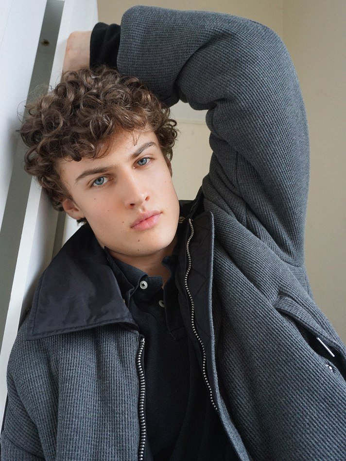 Albert, Montréal male model signed with MTL Models MGMT with curly hair and blue eyes leaning against a white railing, wearing a gray jacket and a black shirt.